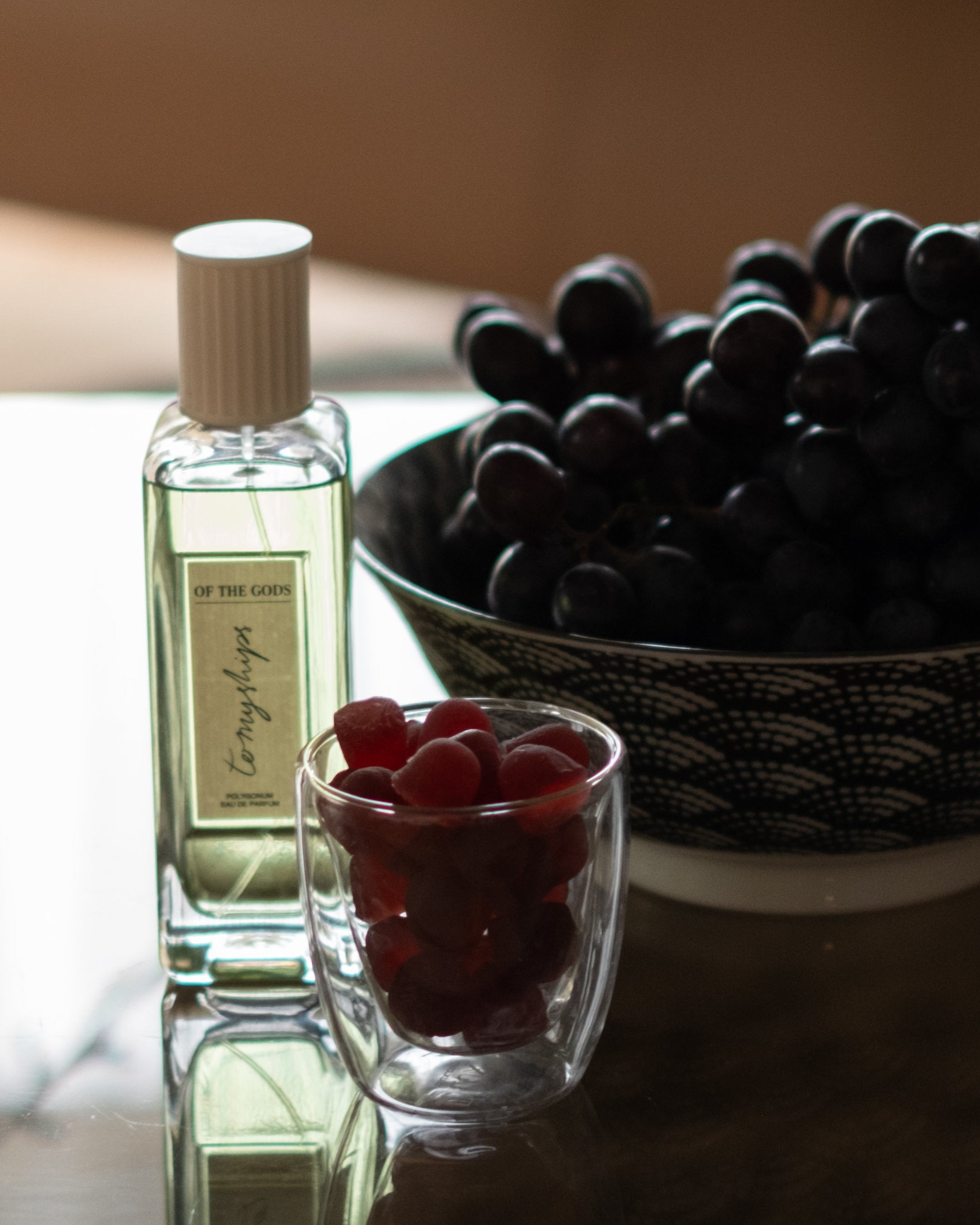 Bottle of perfume, glass with berries, and bowl of grapes on a reflective surface.