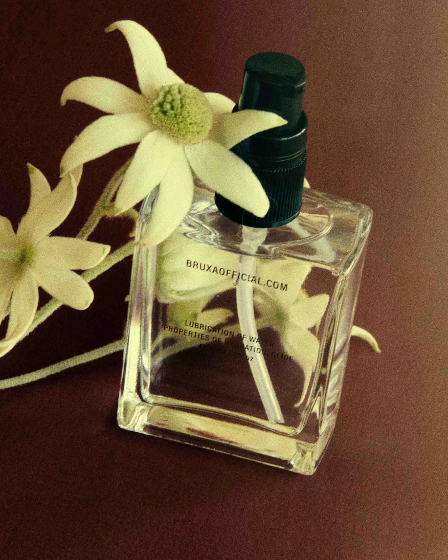 This image features an elegantly designed bottle of BRUXA body oil, positioned prominently in the center. The bottle is made of clear glass and is fitted with a black pump, making it both stylish and functional.