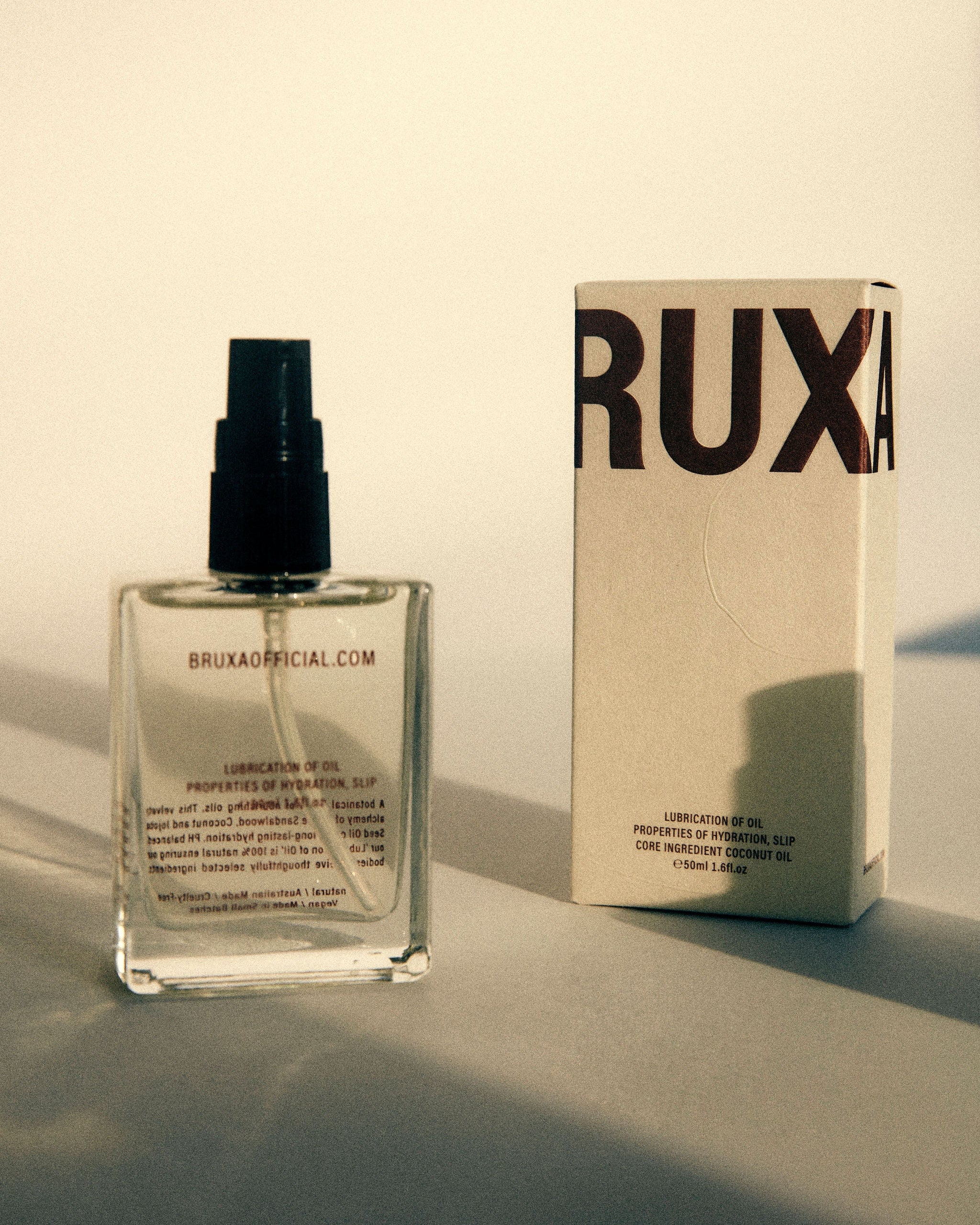 Clear spray bottle with black cap and 'RUXA' branding next to a cardboard box on a light background.
