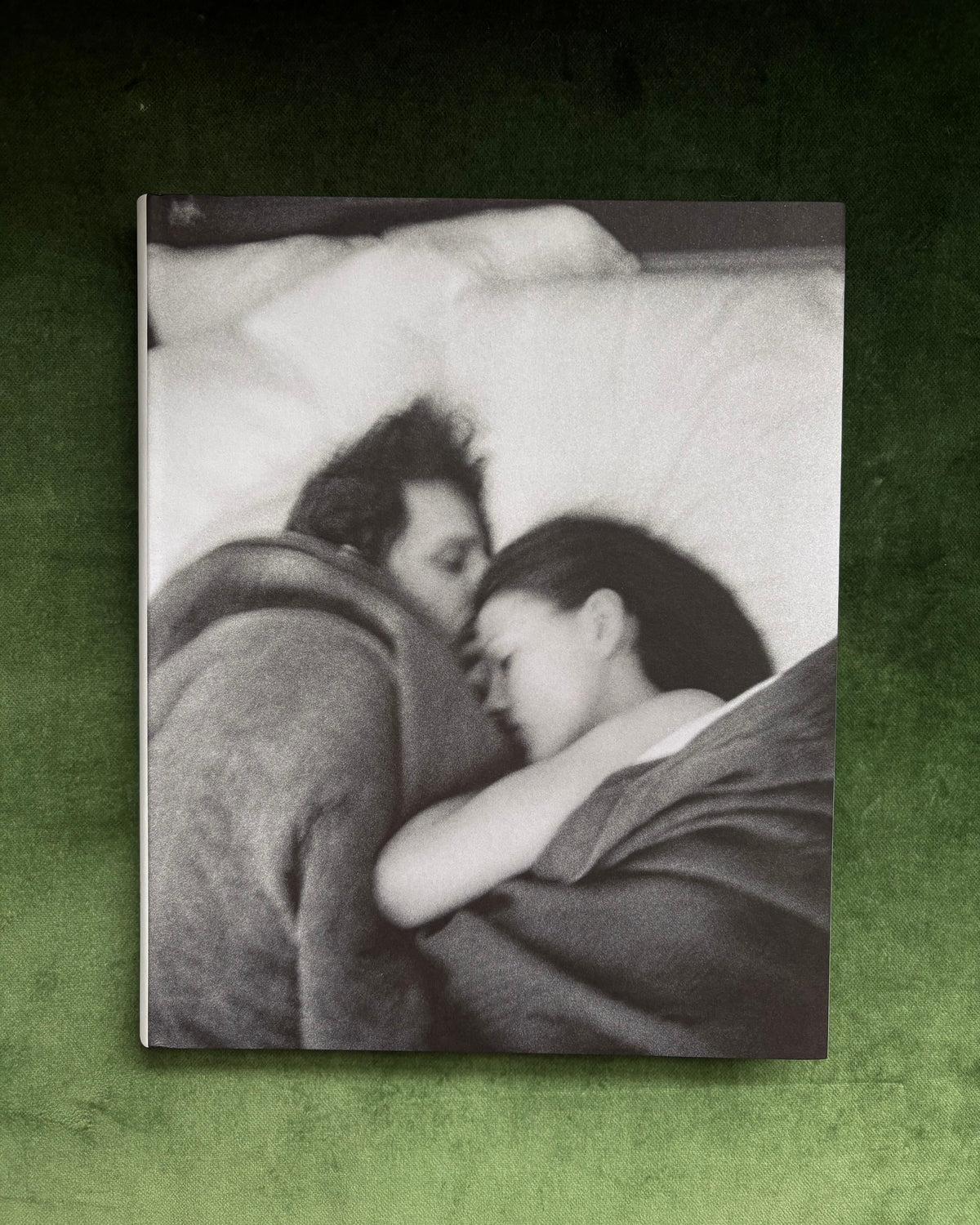 A square black-and-white photographic print or book cover featuring a couple embracing in bed, displayed against a textured dark green background.
