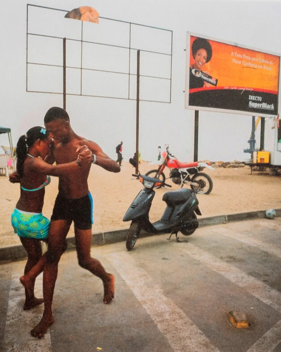 A shirtless man and a woman dancing in a street setting, with a scooter, a dirt bike, and a large outdoor billboard in the background.