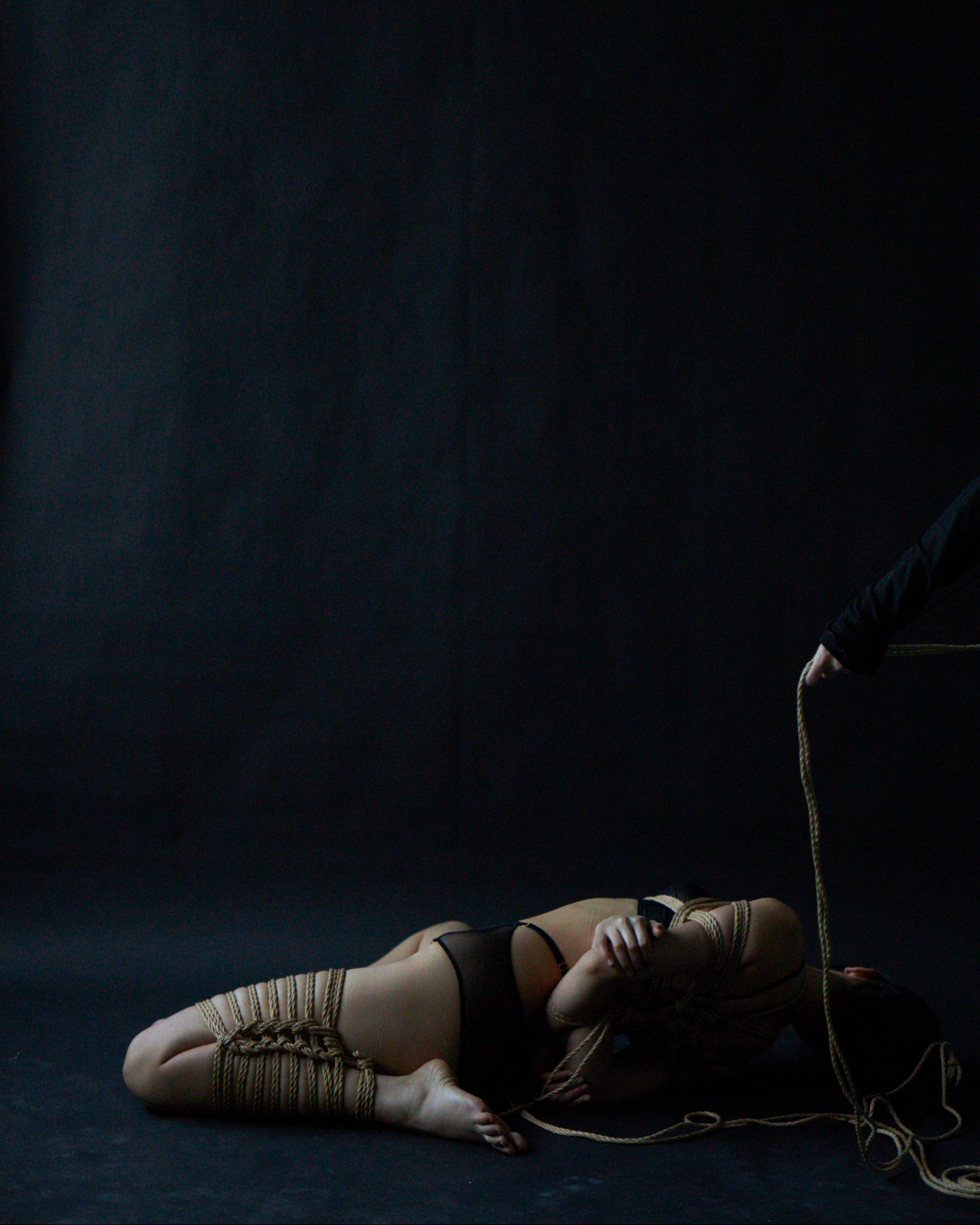 Person lying on a dark surface with a rope, against a black background
