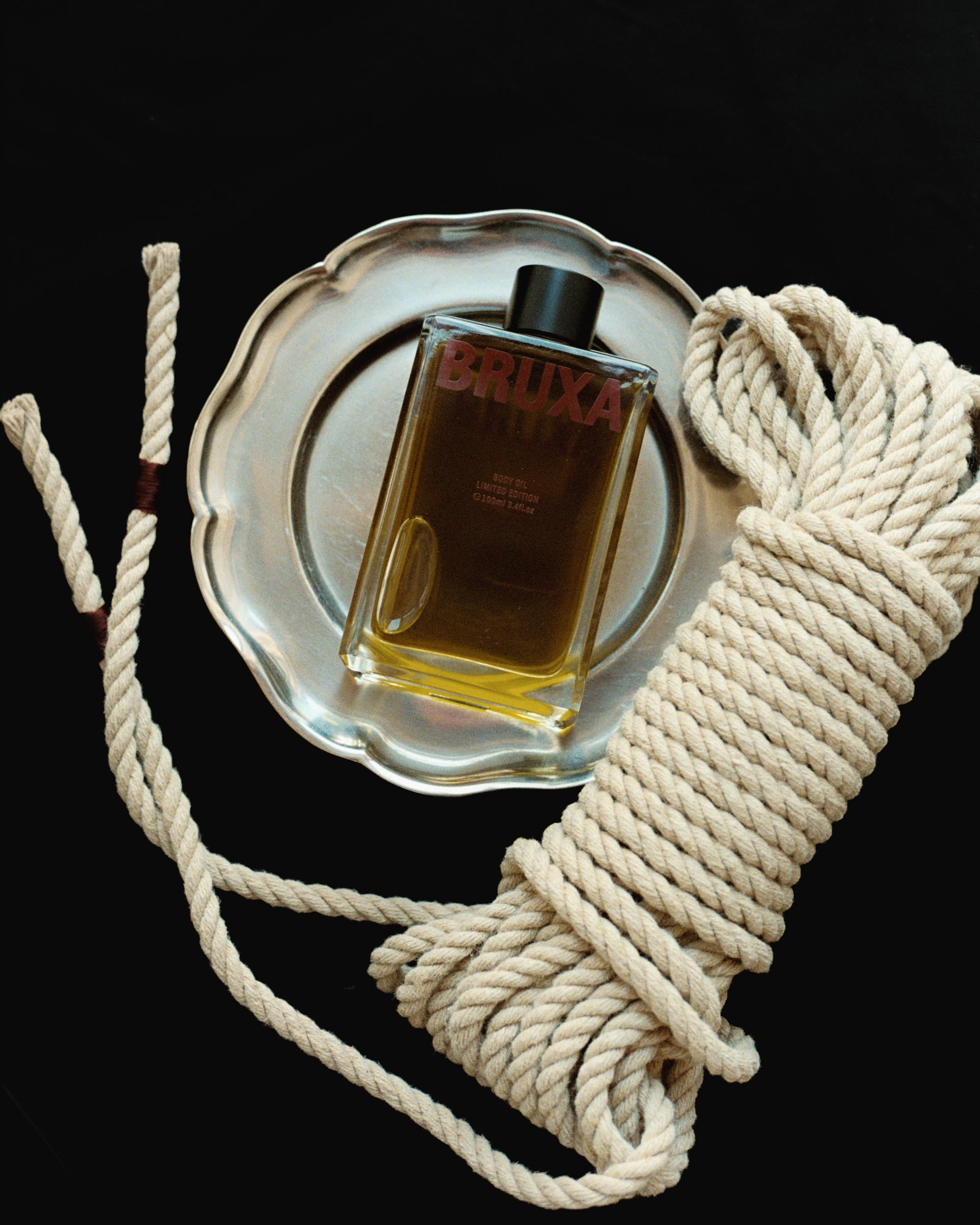 Bottle of Bruxa body oil on a silver tray with a coiled rope on a black background
