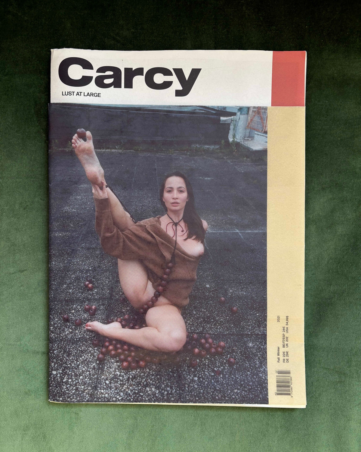 A vintage magazine titled 'Carcy' laid flat on a dark green textured fabric background. The cover features a photograph of a woman in a brown outfit sitting on asphalt with grapes scattered around her, with a cream and red header.