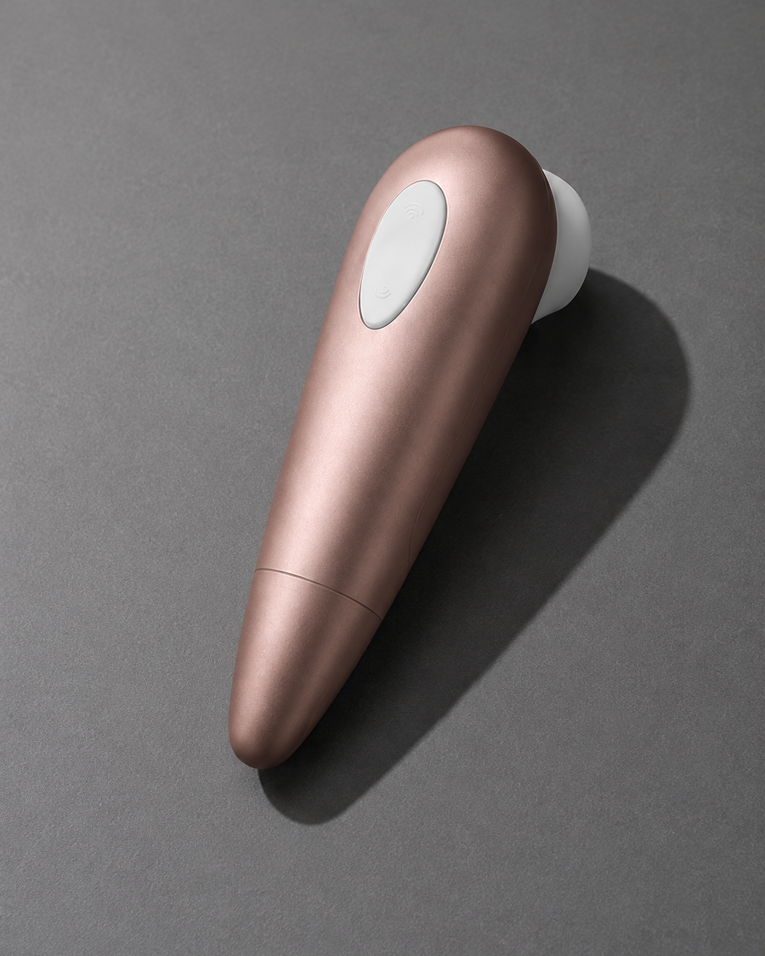 A sleek, metallic rose gold personal massager with a white circular button and white tip, resting on a dark gray surface with a soft shadow.