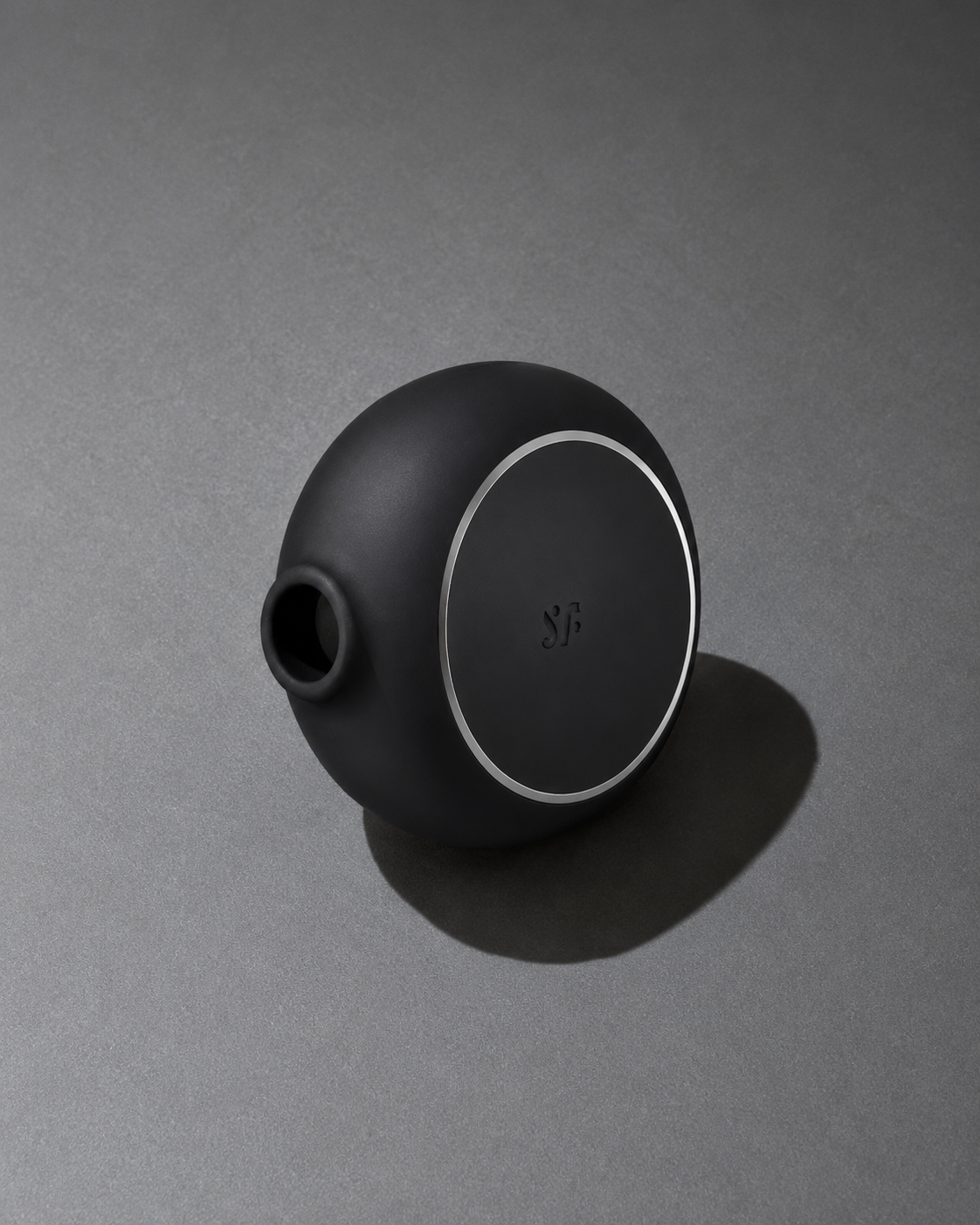A matte black spherical vessel with a small spout and a circular silver ring detail, resting on a dark gray surface with a soft shadow.